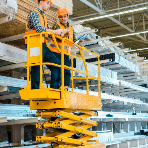 multicultural warehouse workers with digital tablet and clipboard standing on scissor lift